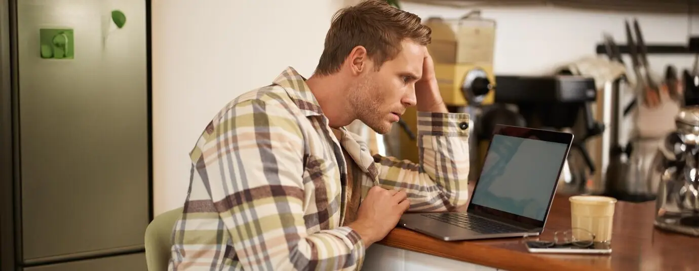 Bild eines erwachsenen Mannes, der in einem Café sitzt, ratlos auf den Laptopbildschirm starrt, online an einer schwierigen Aufgabe arbeitet und dabei Kaffee trinkt.